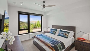 Quarto moderno com uma cama grande, um ventilador de teto, uma mesa de cabeceira e vista para uma paisagem verde através de uma janela ampla.
