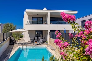 Ein modernes Haus mit zwei Etagen, einem Swimmingpool, Gartenmöbeln und rosa Blumen.