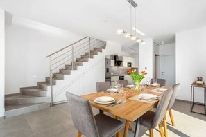 Un espace repas moderne avec une table en bois dressée pour quatre personnes, des chaises à revêtement gris et un escalier avec des rampes en métal menant à un niveau supérieur.