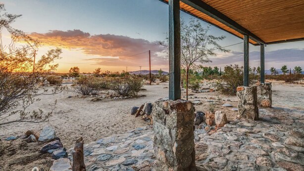 Un espacio al aire libre cubierto, con pilares de piedra y techo de madera, que da vista a un paisaje desértico con vegetación escasa y colinas lejanas.