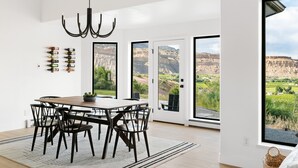 A dining area with a wooden table and chairs, a chandelier, and a view of a mountainous landscape through large glass doors. - The Mesa ViewHouse – Private Pool & Stunning Views – 3BR/3.5BA (Palisade)