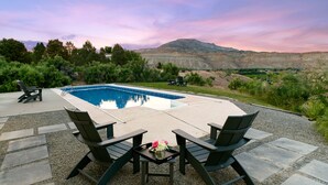 A poolside area with a table and chairs, surrounded by a gravel patio, and a view of a mountainous landscape. - The Mesa ViewHouse – Private Pool & Stunning Views – 3BR/3.5BA (Palisade)