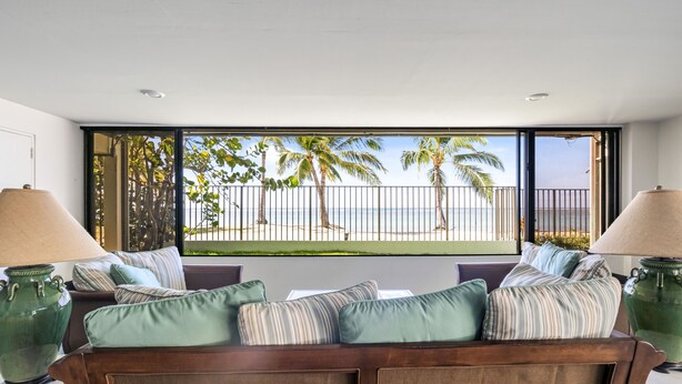 Ein Zimmer mit großem Fenster und Blick auf den Strand und Palmen. Ein Sofa mit gestreiften Kissen und zwei große Vasen auf beiden Seiten.