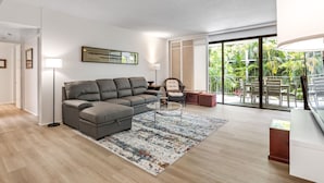 Ein modernes Wohnzimmer mit einem grauen Ecksofa, einem Glastisch und einem großen Fenster mit Blick auf Grünflächen.