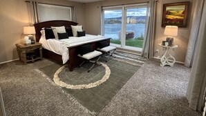 Une chambre à coucher comprenant un lit, un bureau, une chaise, une table de chevet, une lampe, un tapis et une fenêtre donnant sur un lac.