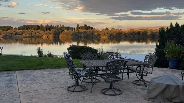 Une terrasse avec une table et des chaises, un barbecue abrité et une vue sur un lac et des arbres.