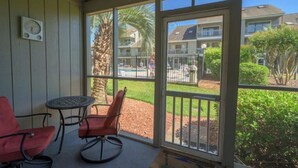 A covered outdoor area with a table and two chairs, a palm tree, and a view of a residential building with a pool. - Poolside Condo in Surfside Beach (Myrtle Beach)
