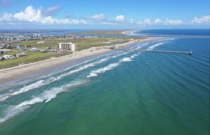 An aerial view of a coastal area with a beach, a pier extending into the water, and a cluster of buildings near the shore. - The Salty Clam (Port Aransas)