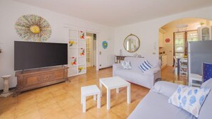 Una sala de estar con televisor, sofá y mesa de centro. Hay una zona de cocina con refrigerador y mesa de comedor.