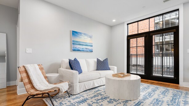Una sala de estar moderna con un sofá blanco, un tapete estampado, una silla de madera y un ventanal con vistas a un edificio de ladrillo.