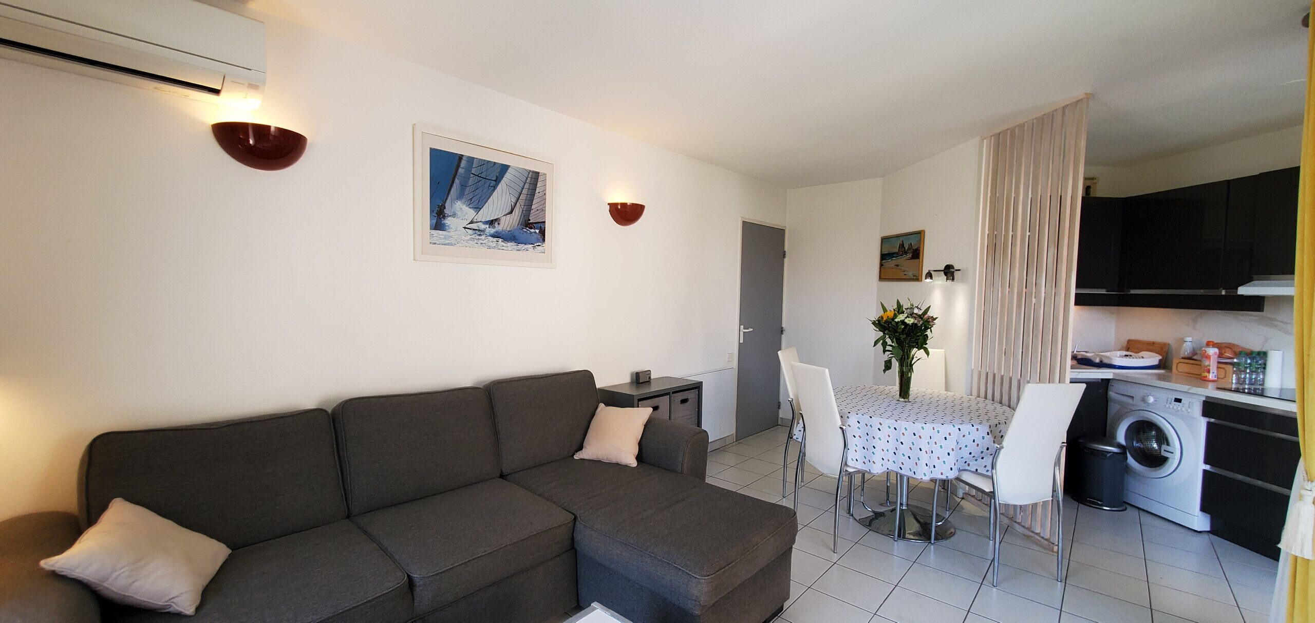 Un espacio habitable compacto con un sofá gris, una mesa de comedor con sillas, una zona de cocina con lavadora, y una pequeña mesa de comedor con un jarrón de flores.