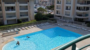 A swimming pool with lounge chairs, a person swimming, and a view of a multi-story building with balconies.