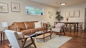 Una sala de estar con un sofá de cuero marrón, un sillón blanco, una mesa de centro de vidrio y una zona de comedor con sillas y una mesa de madera.