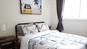 Un dormitorio con cama, mesita de noche, lámpara, ventana con cortinas y un cuadro con motivos playeros en la pared.