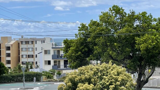 A residential area with multi-story buildings, a street with traffic signs, and a large tree with dense foliage. - Modern Kings Beach: Walk to Beach (CALOUNDRA)