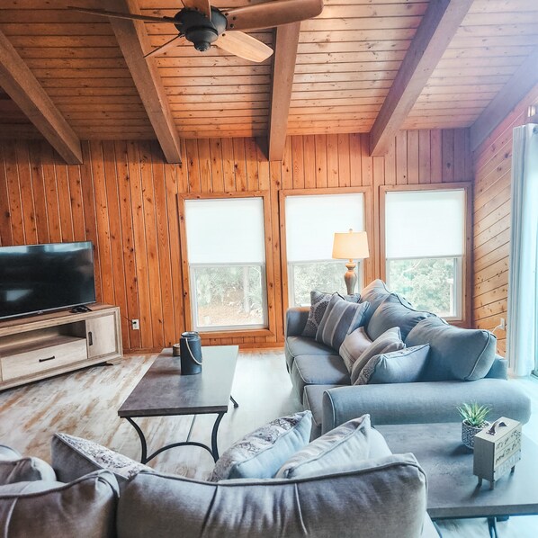 A wooden-paneled room with a large TV, a sofa, a coffee table, and two windows with blinds. - Cozy Island Getaway (2br/2ba)- Pet friendly! Walk to Beach -Community Pool (Eastpoint)