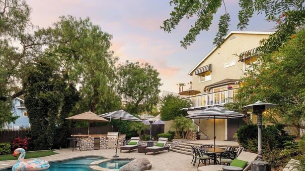 A swimming pool with lounge chairs and umbrellas, surrounded by trees and a building with a balcony. - 5br Home with Pool, Hot Tub, BBQ, Putting green (La Mesa)