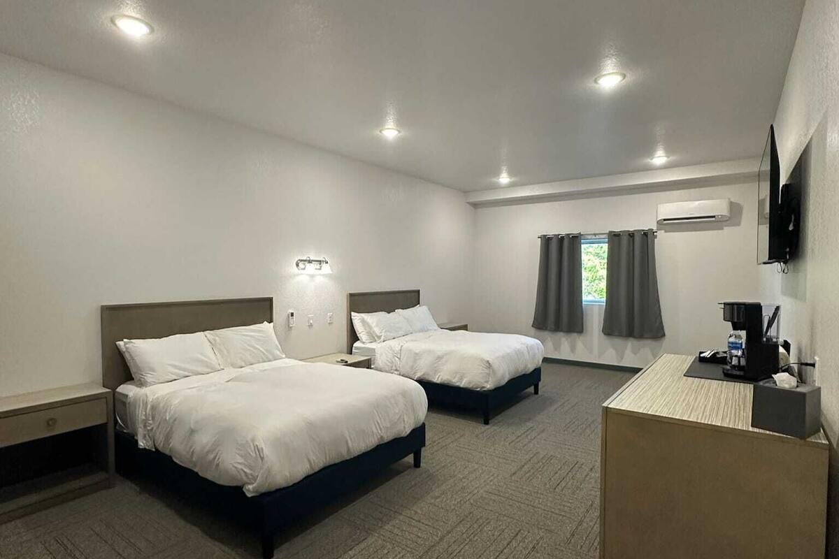 A hotel room with two beds, a desk, a flat-screen TV, and a window with curtains.