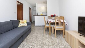 A compact living space with a grey sofa, a dining table with chairs, a kitchen area, and a TV on a wooden stand. - Peñismar Garden by Interhome (Peñiscola)