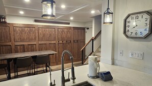 Una cocina moderna con una isla grande, pileta de acero inoxidable, paneles de madera, iluminación colgante y un reloj de pared.