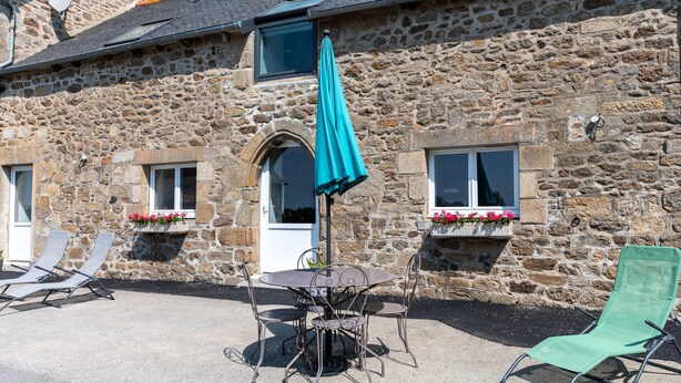 A stone building with a patio area featuring a table and chairs, a green umbrella, and a green lounge chair. - Gîtes Ti Tud-kozh, Location "L' Arguenon" (PLUDUNO)
