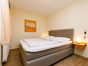 Un lit bien fait, avec une tête de lit grise, une table de chevet sur laquelle se trouve une lampe, et une œuvre d’art accrochée au mur.