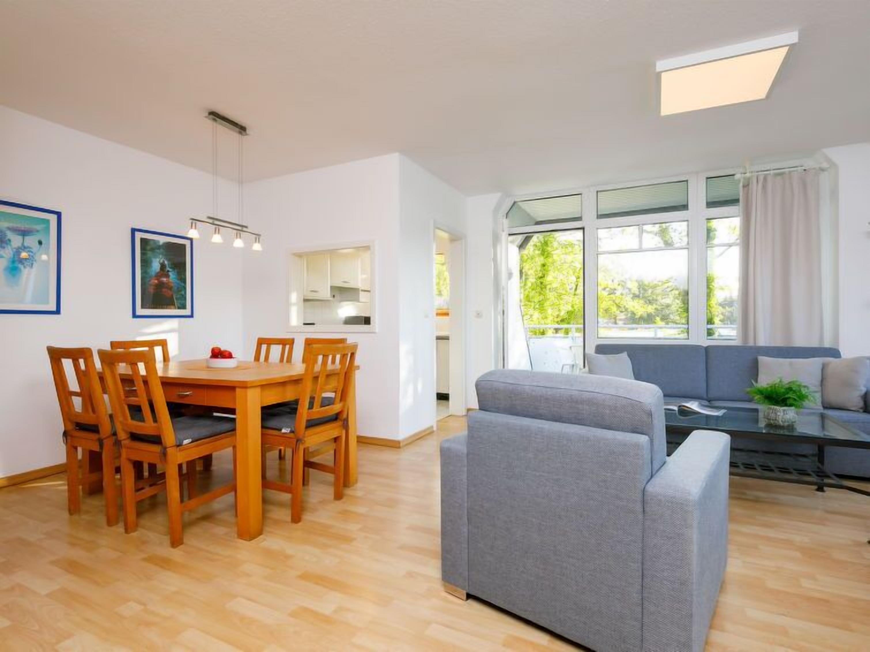 A modern living room with a grey sofa, wooden dining table, and chairs. There is a large window with curtains, a ceiling light, and a view of greenery outside.