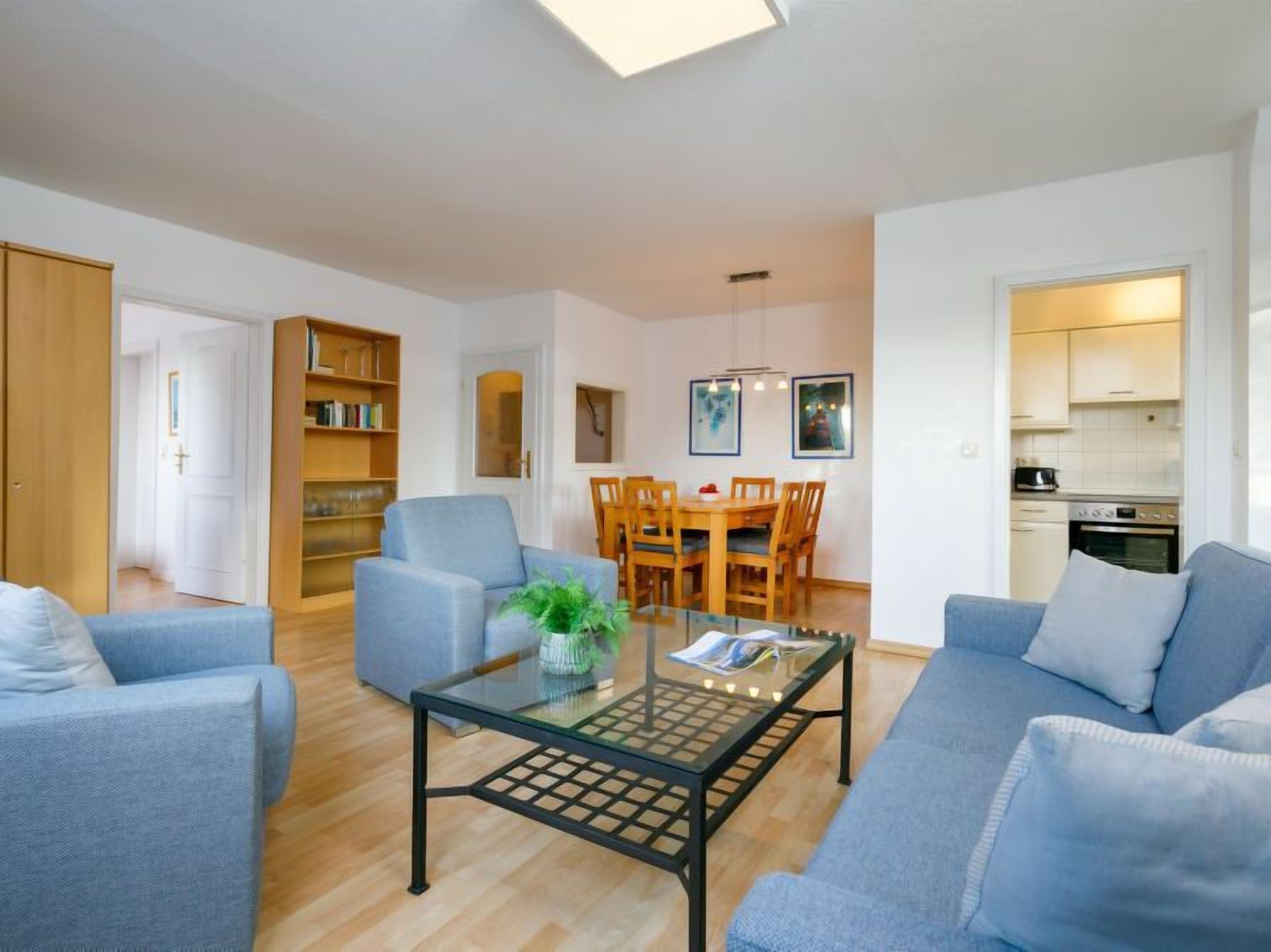 Ein modernes Wohnzimmer mit einem Glastisch, blauen Sofas, einem Essbereich mit Holzsesseln und einer durch eine offene Tür sichtbaren Küche.