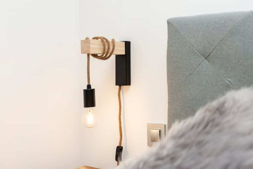 Una lámpara de pared con cable de madera y un enchufe negro, un cabecero tapizado gris y un enchufe eléctrico visible.