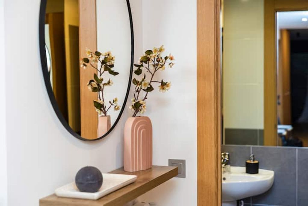 Un baño con un espejo redondo, un jarrón rosa con flores y un lavabo blanco.