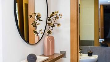 Un baño con un espejo redondo, un jarrón rosa con flores y un lavabo blanco.