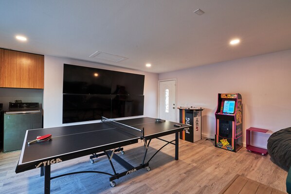 A room with a ping pong table, a small kitchenette, a large window, and a colorful arcade machine.