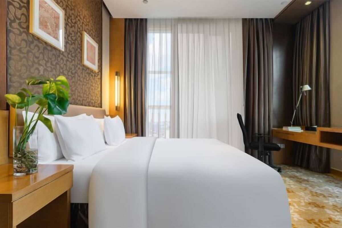 Ein Hotelzimmer mit einem großen Bett, einem Schreibtisch mit Stuhl, einer Pflanze und einem Fenster mit Vorhängen.