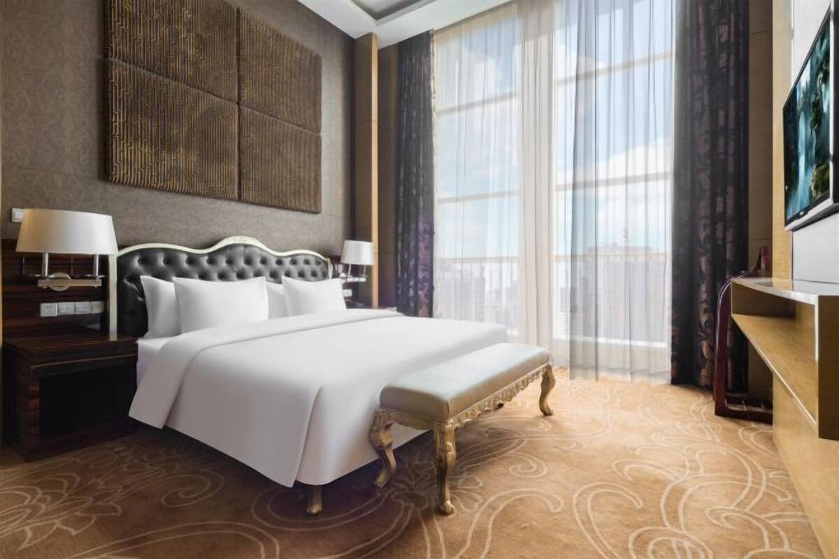 Ein Hotelzimmer mit einem großen Bett, einem Nachttisch, einem Schreibtisch und einem großen Fenster mit Vorhängen.