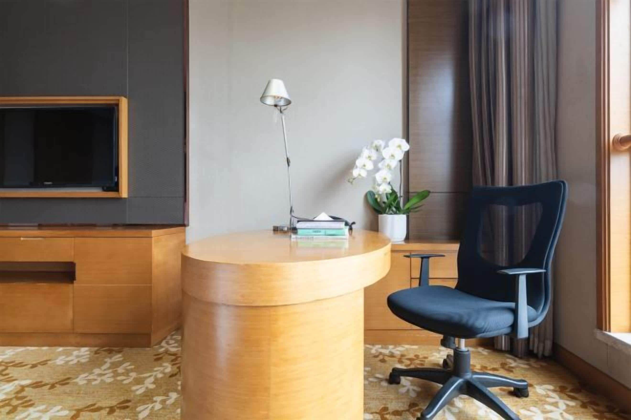 A hotel room with a wooden desk, a chair, a TV, a lamp, a potted plant, and a window with curtains.