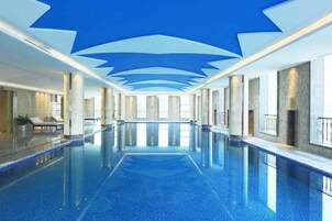 Una amplia piscina interior con suelo azul cristalino, rodeada de muros y columnas, y un techo con un diseño geométrico.