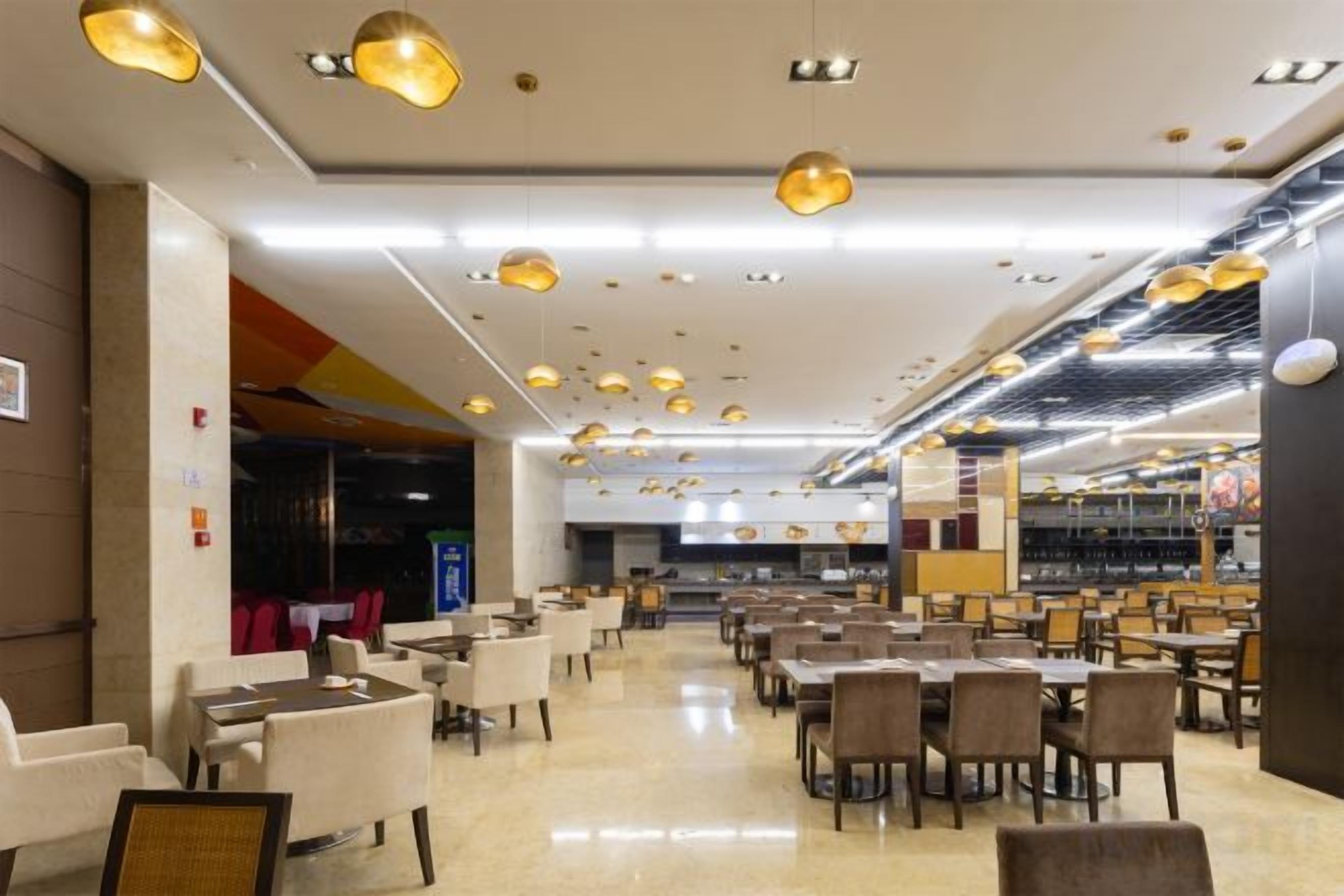 A spacious indoor area with multiple tables and chairs, ceiling lights, and a visible kitchen area in the background.