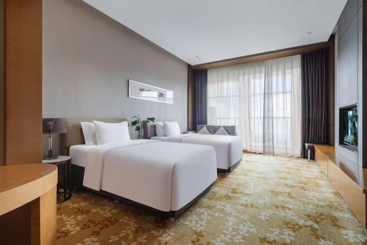 Ein Hotelzimmer mit zwei Betten, einem Fernseher, einem Schreibtisch und einem großen Fenster mit Vorhängen.
