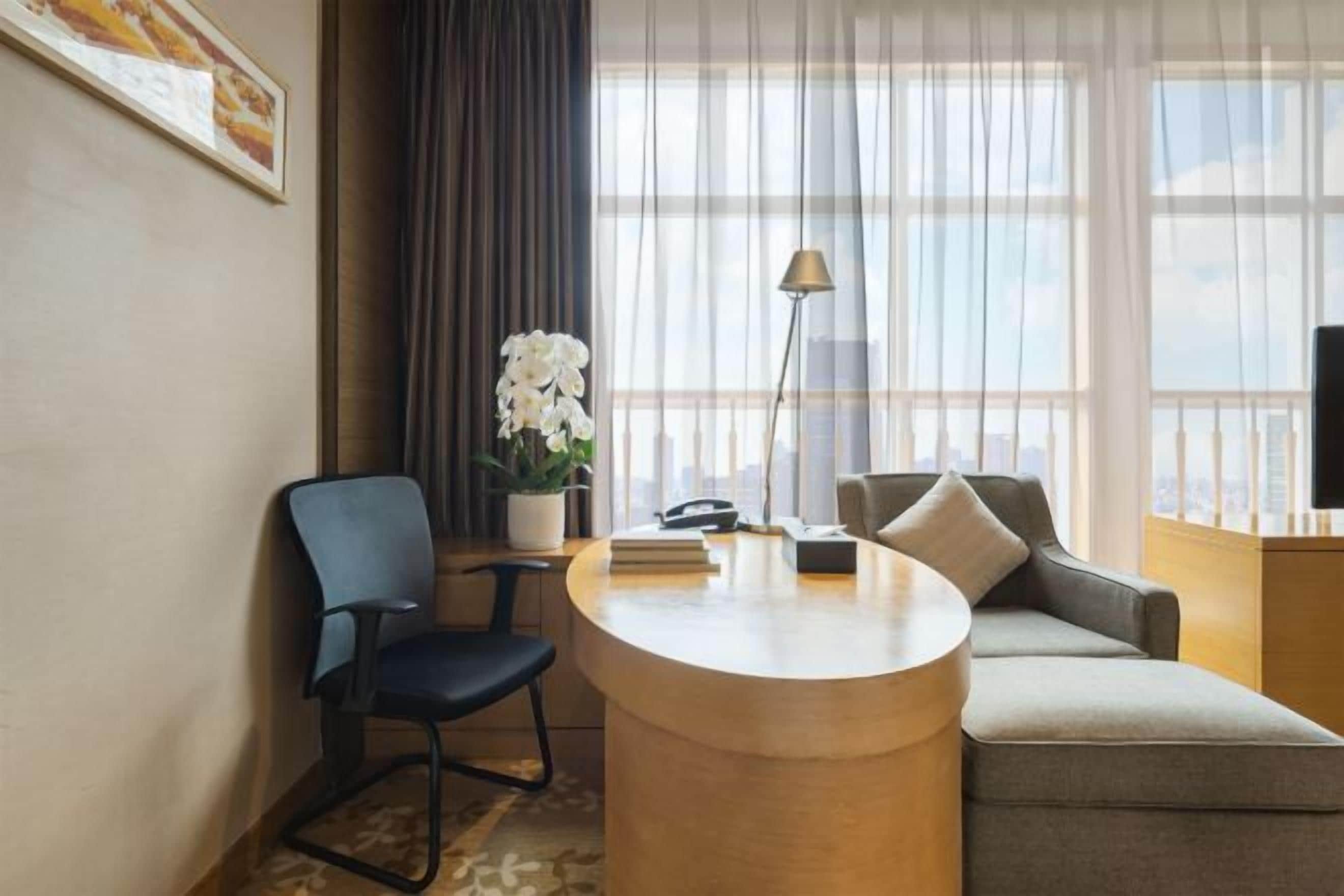 A hotel room with a round wooden desk, a blue chair, a grey sofa, a potted plant, a lamp, and a television.