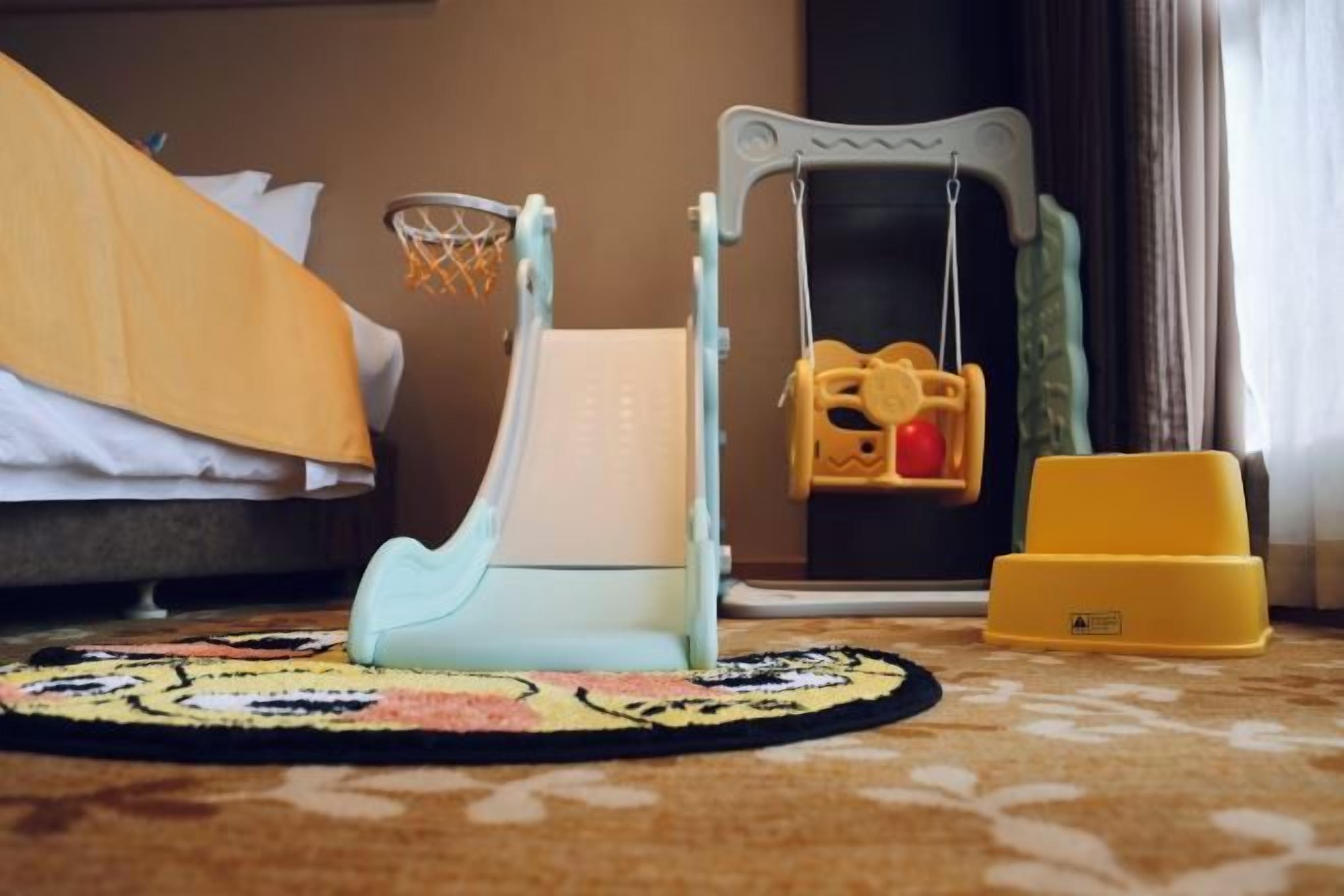 Una habitación de hotel con una cama, un pequeño espacio de juegos con un columpio y un caja de almacenamiento amarilla.