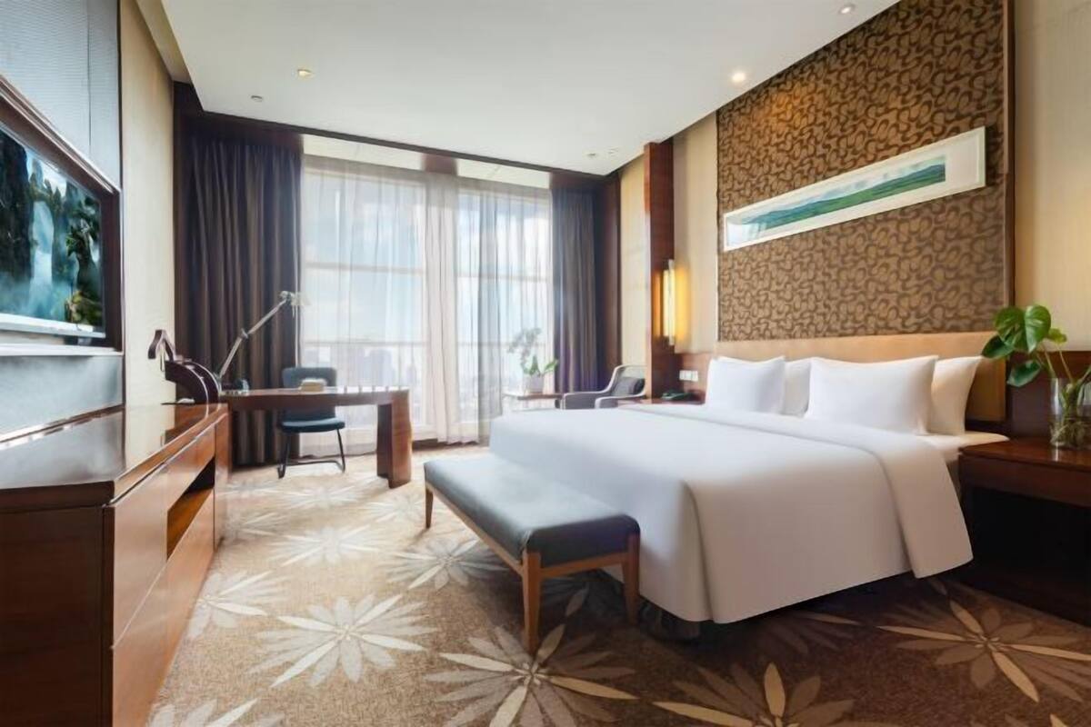 Ein Hotelzimmer mit einem großen Bett, einem Schreibtisch, einem Sessel, einem Fernseher und einem Fenster mit Vorhängen.