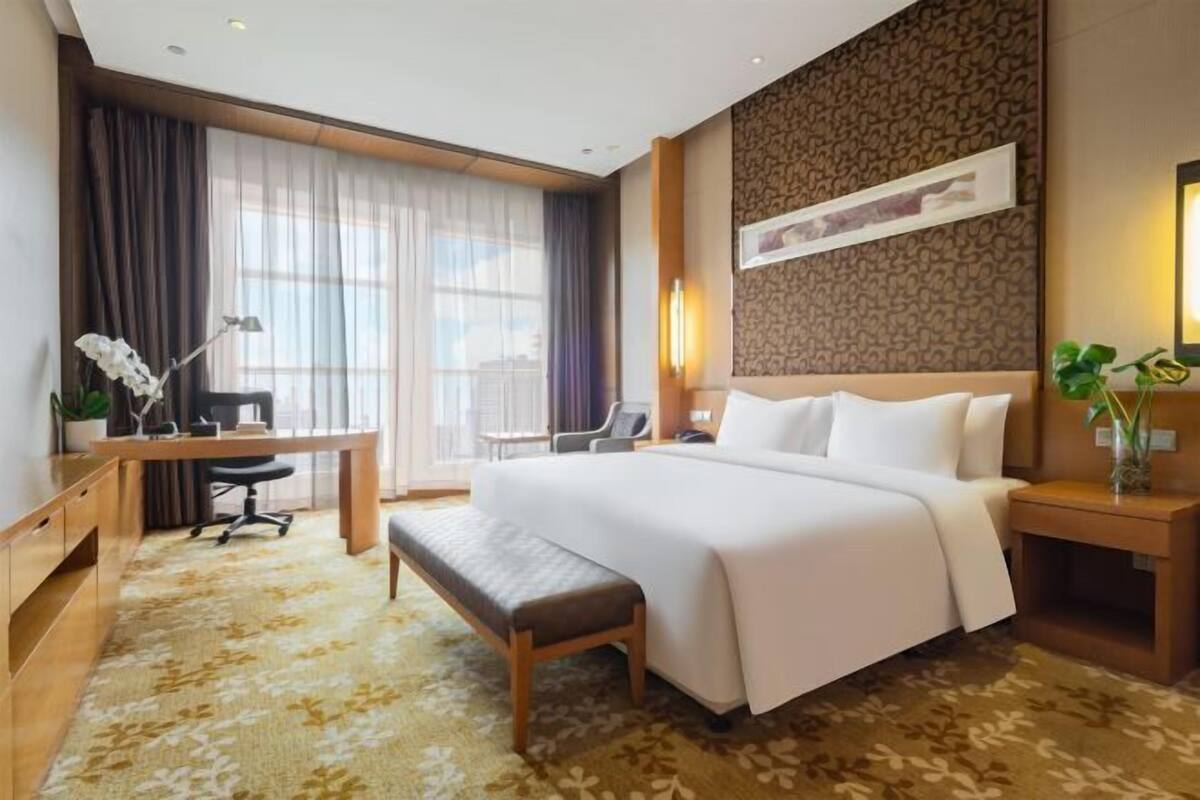 Ein Hotelzimmer mit einem großen Bett, einem Schreibtisch mit Stuhl, einer Bank, einer Schreibtischlampe, einem Nachttisch mit Lampe und einem Fenster mit Vorhängen.