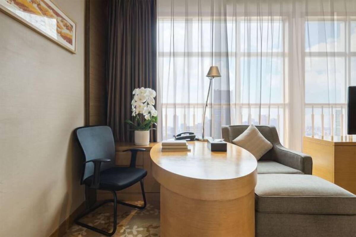 Ein Hotelzimmer mit einem runden Holztisch, einem blauen Stuhl, einem grauen Sofa, einer Topfpflanze, einer Lampe und einem Fernseher.