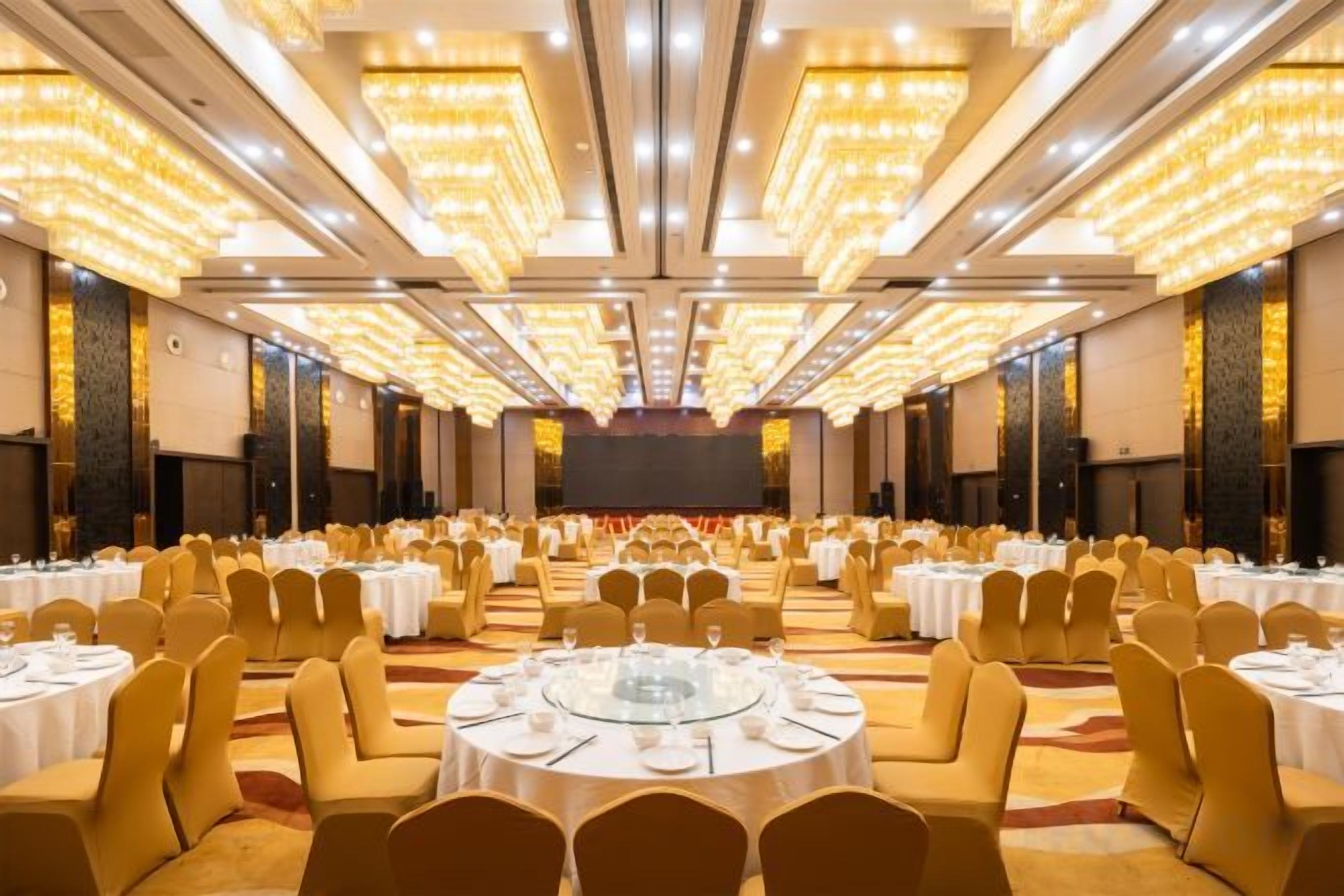 Un salón de banquetes con mesas y sillas redondas, un escenario al frente y un techo alto con luces.