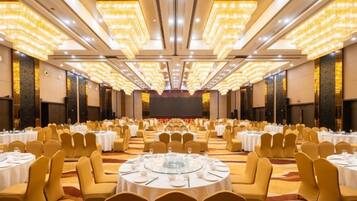 Un salón de banquetes con mesas y sillas redondas, un escenario al frente y un techo alto con luces.