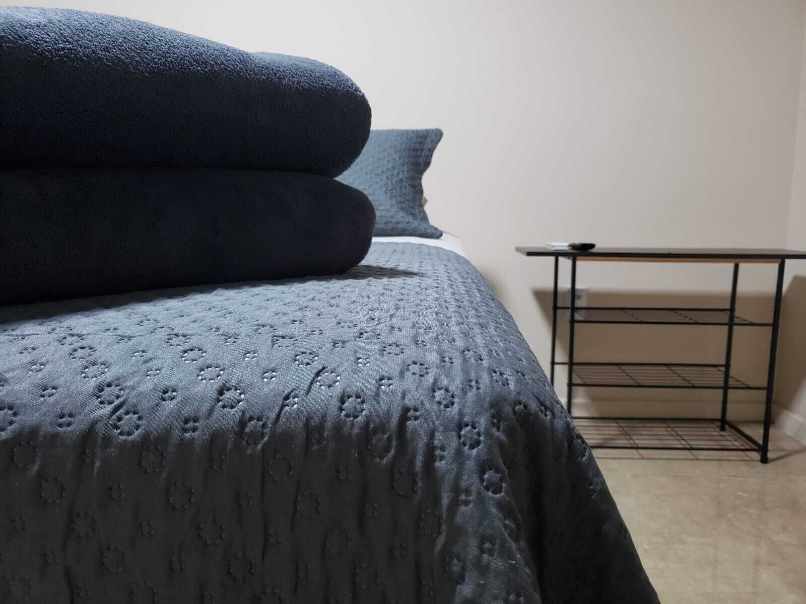 A neatly made bed with a patterned bedspread and pillows, a small black metal table beside it, and a plain wall in the background.