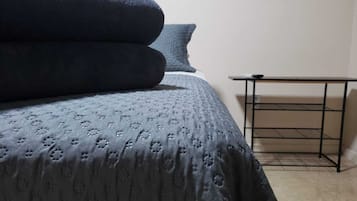 A neatly made bed with a patterned bedspread and pillows, a small black metal table beside it, and a plain wall in the background.
