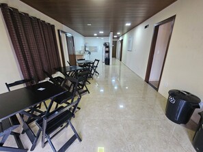 A hotel corridor with a dining area featuring tables and chairs, a trash bin, and a kitchen area in the background. - Hospedagem Costa BNU (Blumenau)