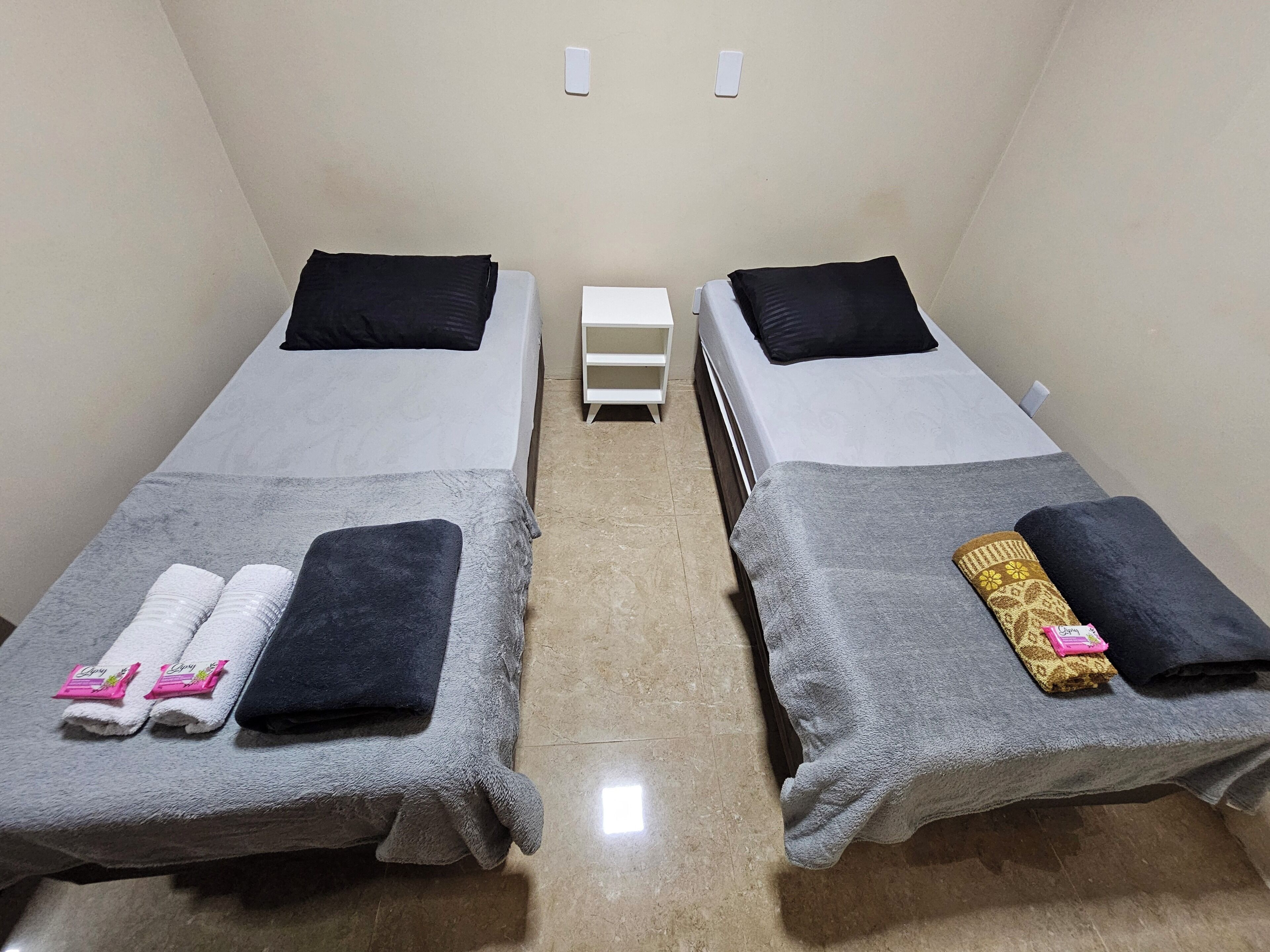 Ein Zimmer mit zwei Betten, jedes mit einem Kopfkissen und einem Handtuch. Dazwischen befindet sich eine kleine, weiße Kommode.