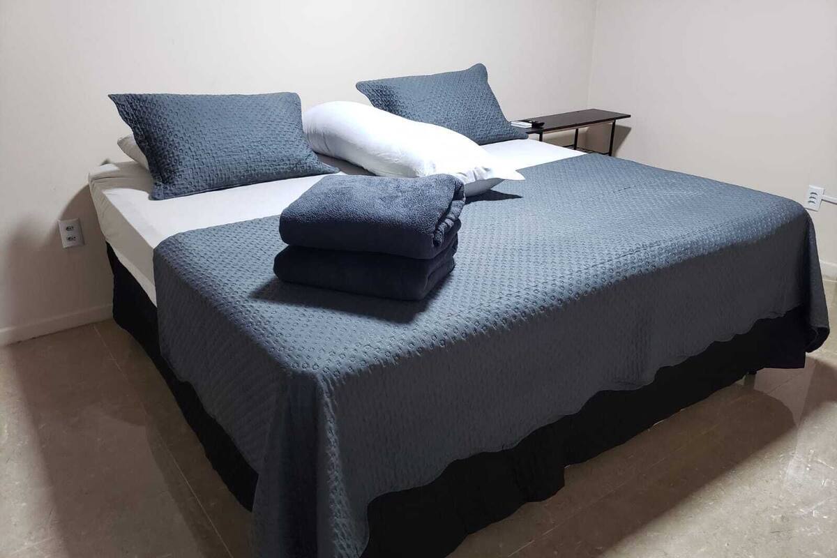 Uma cama bem arrumada com colcha azul escura e almofadas, um colchão branco e uma pequena mesa de cabeceira com abajur.